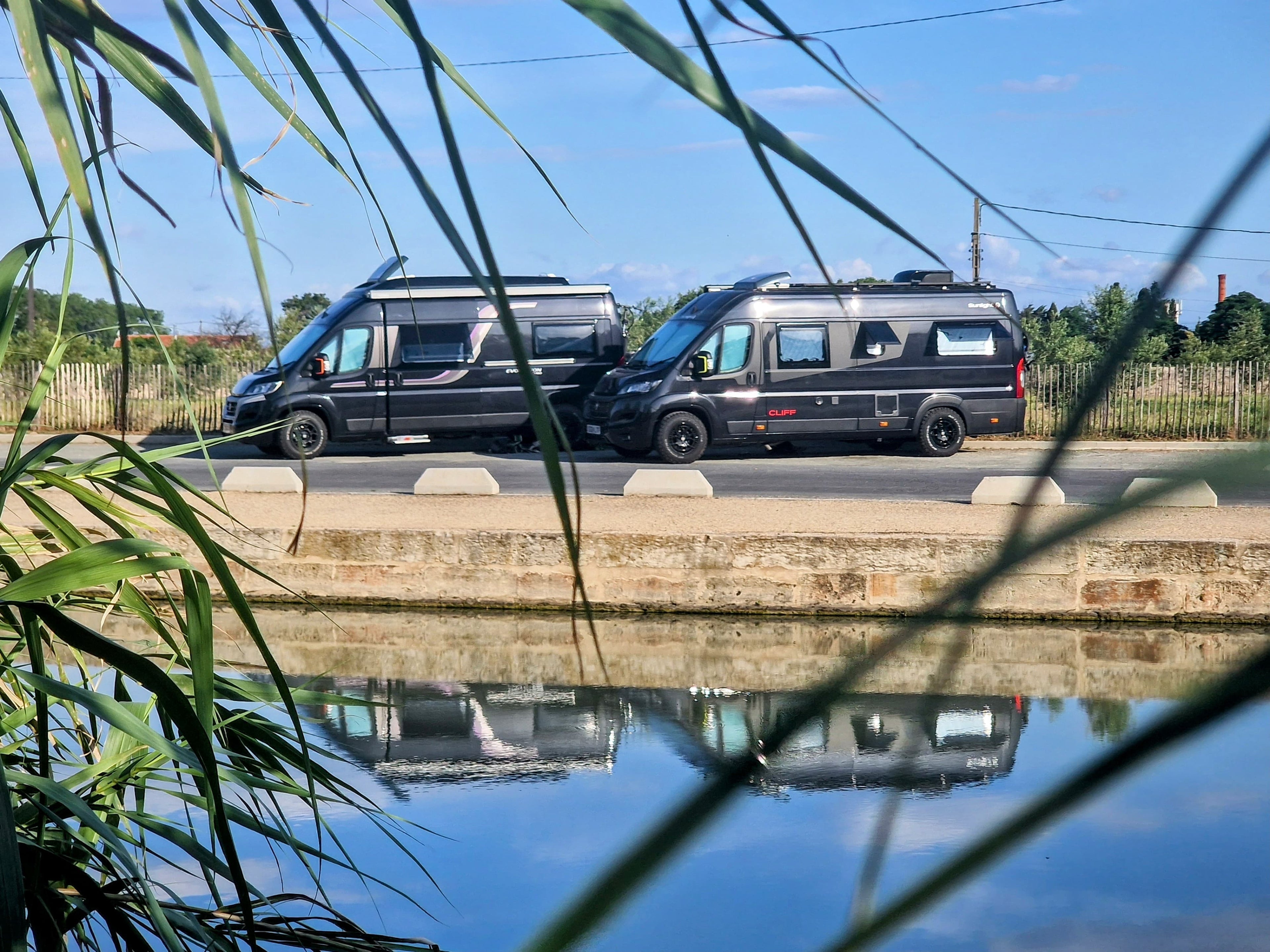 Two black vans parked on a road with a reflective water surface and greenery in the foreground.